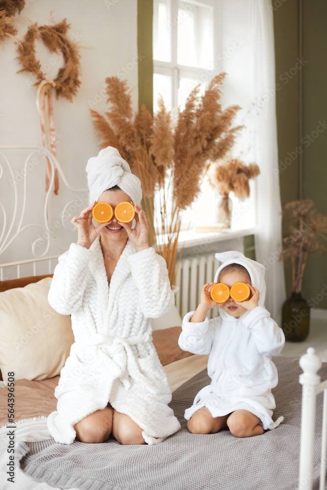 Full body faceless portrait of beautiful mother and cute little daughter having fun, sitting on bed with fresh fruit orange for face mask having spa day toghether at home - obrazy, fototapety, plakaty 