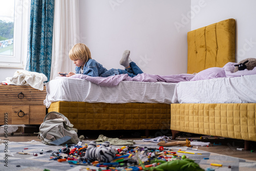Obraz na płótnie boy using phone, playing phone games in messy childrens room, kid playing among