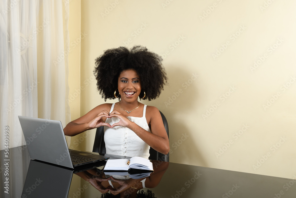 woman smiles and makes a heart shape with her hands at her workplaceon the table a laptop, young Afro-Brazilian woman woman smiles and makes a heart shape with her hands at her workplaceon the table a laptop, young Afro-Brazilian woman