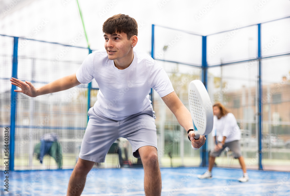 Concentrated young guy paddle tennis player performing left-handed forehand to return ball on outdoor court on sunny summer day..