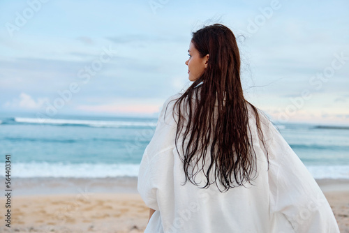 Happy tanned slender woman in white swimsuit shirt and denim shorts walks on the beach with her back to the camera on the sand by the ocean with wet hair after swimming, sunset light