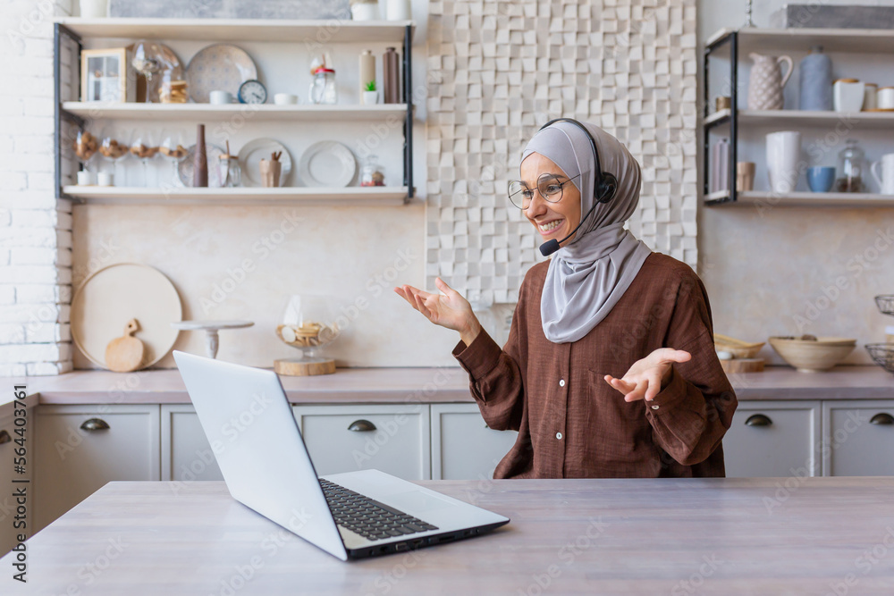 Joyful woman in hijab working from home remotely, Muslim woman talking on video call using headset and laptop, businesswoman working remotely at home.