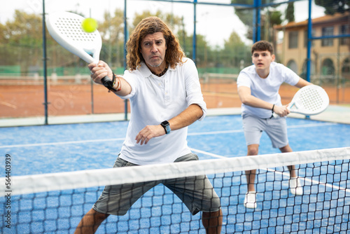 Sportive men of different ages padel players hitting ball with racket on hard court in summer © JackF
