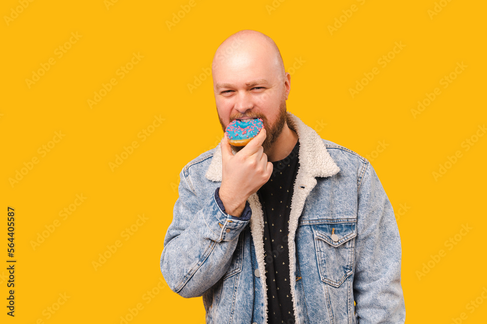 Young bald bearded man wearing jeans jacket bites from a blue doughnut. Young bald bearded man wearing jeans jacket bites from a blue doughnut.
