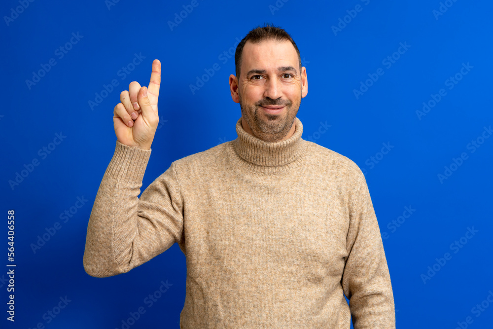 Naklejka premium Insightful intelligent proactive man hispanic man 40s wearing turtleneck holding index finger up with great new idea isolated on blue background studio portrait. People lifestyle concept.