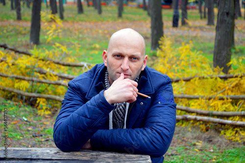 bald young man in a jacket smokes a cigarette on a bench in the autumn park. a man enjoys a calm rest, fragrant cigarette smoke and a good autumn day