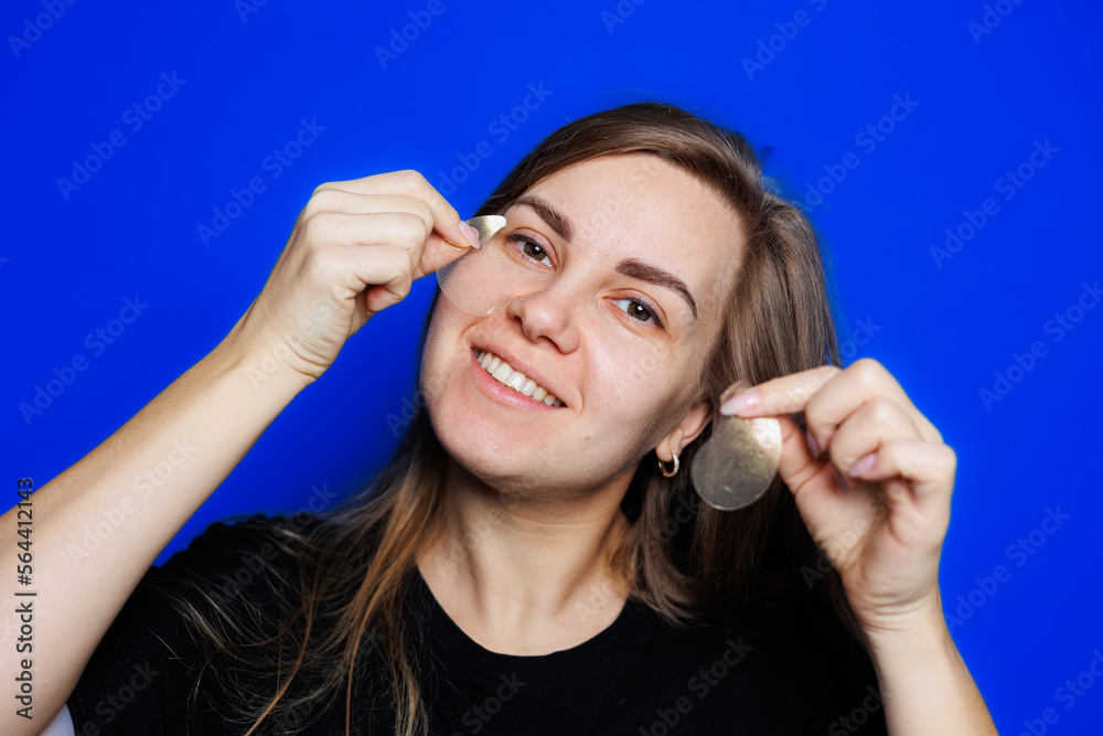 Cute young blonde woman with natural smooth skin without makeup posing happy and smiling at camera while standing in t-shirt on blue background. Beauty concept, eye patches for women Cute young blonde woman with natural smooth skin without makeup posing happy and smiling at camera while standing in t-shirt on blue background. Beauty concept, eye patches for women