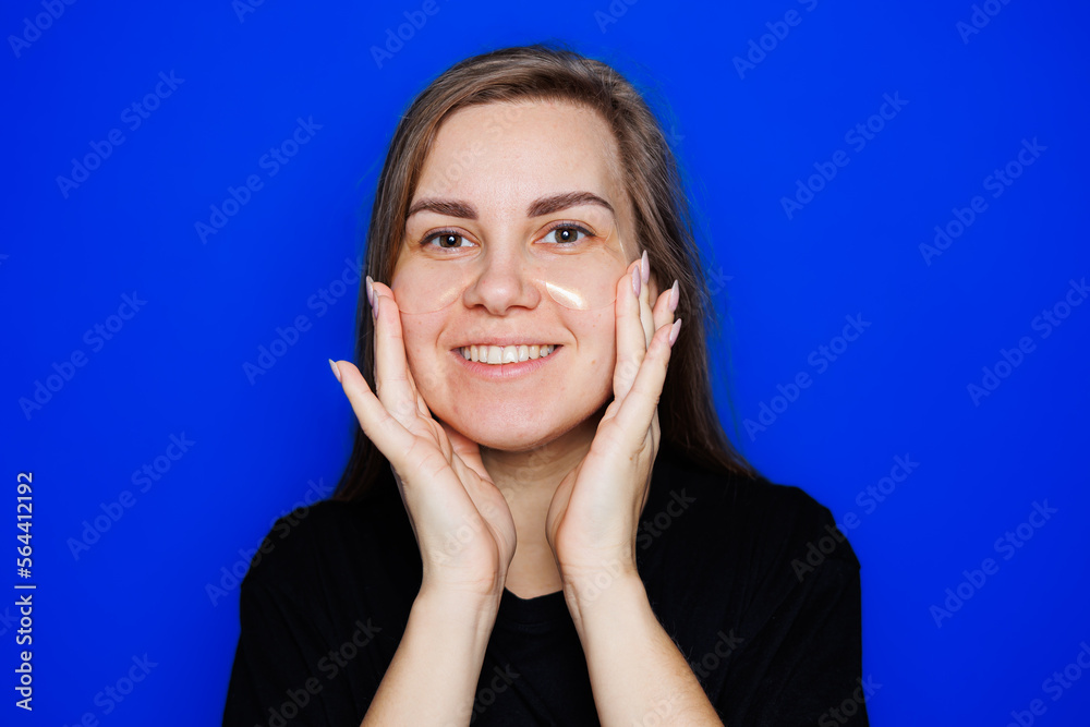 Cute young blonde woman with natural smooth skin without makeup posing happy and smiling at camera while standing in t-shirt on blue background. Beauty concept, eye patches for women Cute young blonde woman with natural smooth skin without makeup posing happy and smiling at camera while standing in t-shirt on blue background. Beauty concept, eye patches for women