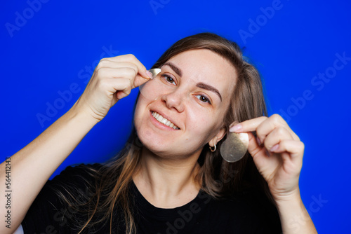 Cute young blonde woman with natural smooth skin without makeup posing happy and smiling at camera while standing in t-shirt on blue background. Beauty concept, eye patches for women