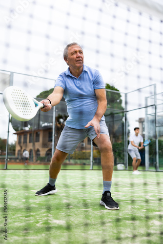 European old man holding playing padel during training in court. View through tennis net