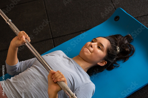 Beautiful sportswoman doing exercises indoors in gym, lying on mat, lifting barbell © pablobenii