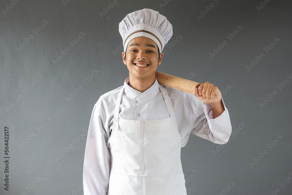 Happy male Asian chef or baker in toque with rolling-pin over grey background Happy male Asian chef or baker in toque with rolling-pin over grey background