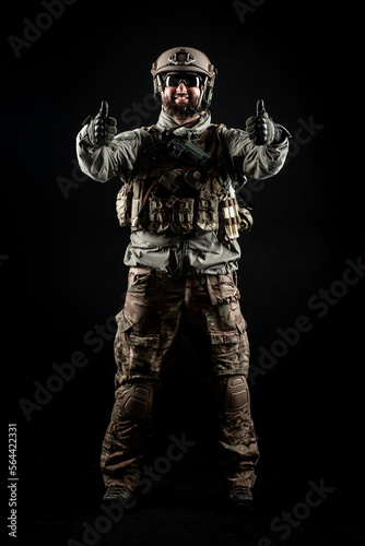 USA soldier in a military suit with a rifle smiles and shows a like against a dark background, American commando in uniform