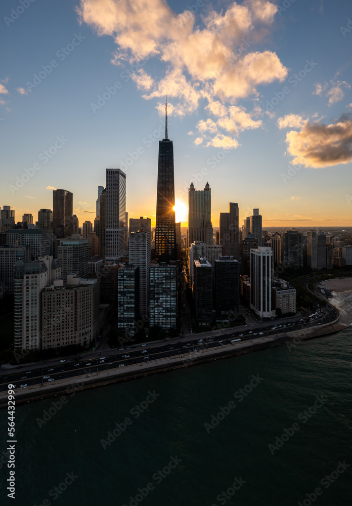 Downtown Chicago aerial as the golden sun peaks out from behind a high-rise building during the autumnal equinox with traffic on Lakeshore Drive along Lake Michigan below.