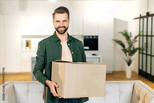 Happy attractive stylish caucasian man, stand at home in living room, holding in hands a large cardboard box, received parcel from the online store, preparing to unpack, looks at camera,smile friendly