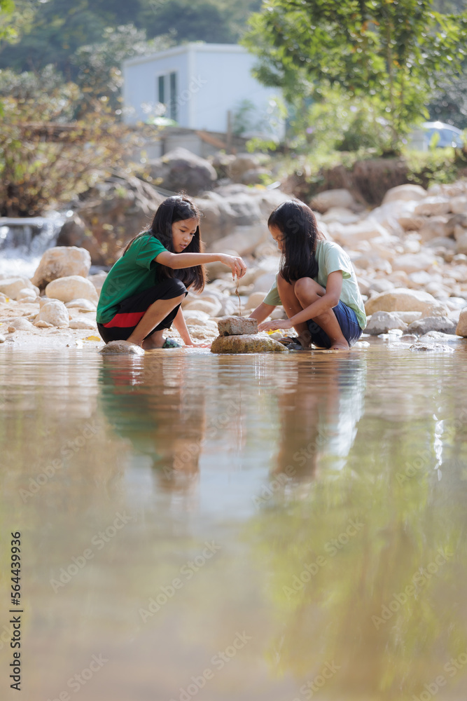 Fototapeta premium Two asian siblings playing sand nearing the river together with fun. Outdoor activity for children on vacation.
