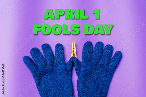 April 1 fools day inscription.A Pair of Knitted blue Gloves on purple Background.The thumb has a Laundry clip.April fool day concept.Copy space.