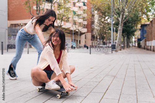 two young women laughing and having a good time playing with a skateboard down a city street, concept of friendship and teenager lifestyle, copy space for text