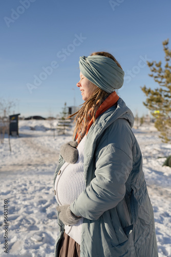 A happy pregnant young woman with her eyes closed, hugging her stomach in the park in winter, in a warm down jacket.