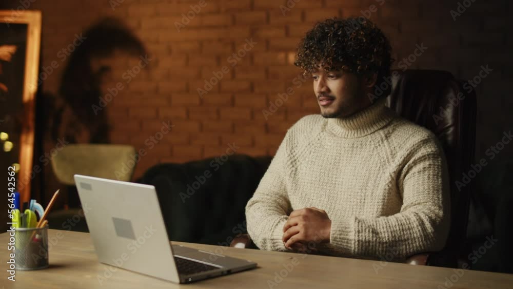Handsome young man in sweater having video call. Curly guy with laptop listening carefully during online conference. Home office, work from home Handsome young man in sweater having video call. Curly guy with laptop listening carefully during online conference. Home office, work from home