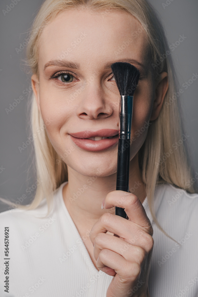 Obraz premium Portrait of a woman covering one eye on the face using makeup brush, looking at camera and smiling isolated over gray.