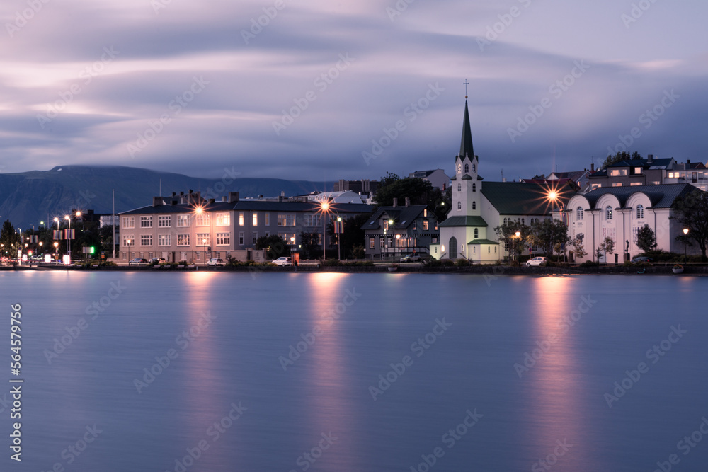 An evening long exposure landscape of Reykjavik city centre with Fríkirkjan church over the Tjörnin lake with blurry clouds on the sky and light reflections on the water