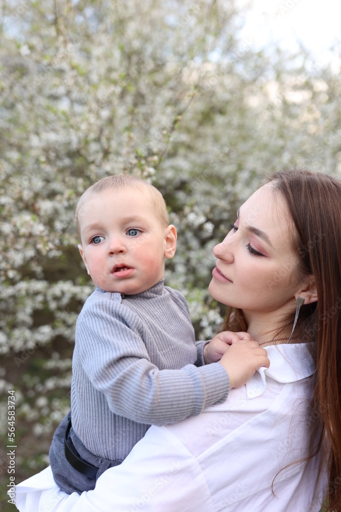 mother hugs her little son in spring near a blooming tree. Spring and plum blossom. Mother's Day. The love of a son and a mother. Gray clothes for a one-year-old boy. Kiss the son. child's laughter mother hugs her little son in spring near a blooming tree. Spring and plum blossom. Mother's Day. The love of a son and a mother. Gray clothes for a one-year-old boy. Kiss the son. child's laughter
