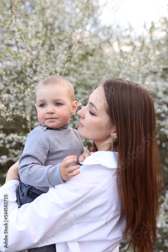 mother hugs her little son in spring near a blooming tree. Spring and plum blossom. Mother's Day. The love of a son and a mother. Gray clothes for a one-year-old boy. Kiss the son. child's laughter