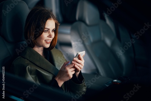 a close horizontal portrait of a stylish, luxurious woman in a leather coat sitting in a black car at night in the passenger seat, looking pleasantly into the camera holding a smartphone in her hand © SHOTPRIME STUDIO