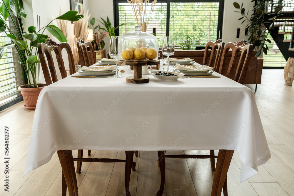 A fully set table with white elegant tablecloth, wooden chairs and food stand containing lemons, standing in the dining room of the house with green plants around ahd blur gardenview on the background