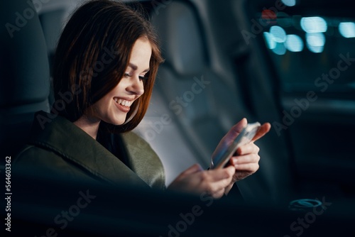 a close horizontal portrait of a stylish, luxurious woman in a leather coat sitting in a black car at night in the passenger seat, happily smiling while looking at her smartphone during the trip