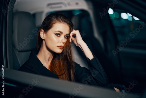horizontal photo from the side, at night, of a woman sitting in a black car and thoughtfully looking at the camera holding her hand near her face, parking the car in the parking lot © SHOTPRIME STUDIO