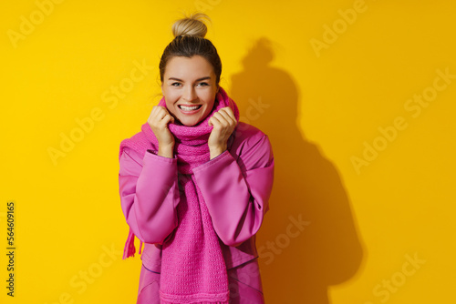 Young cheerful woman wearing pink coat and wool scarf against yellow background