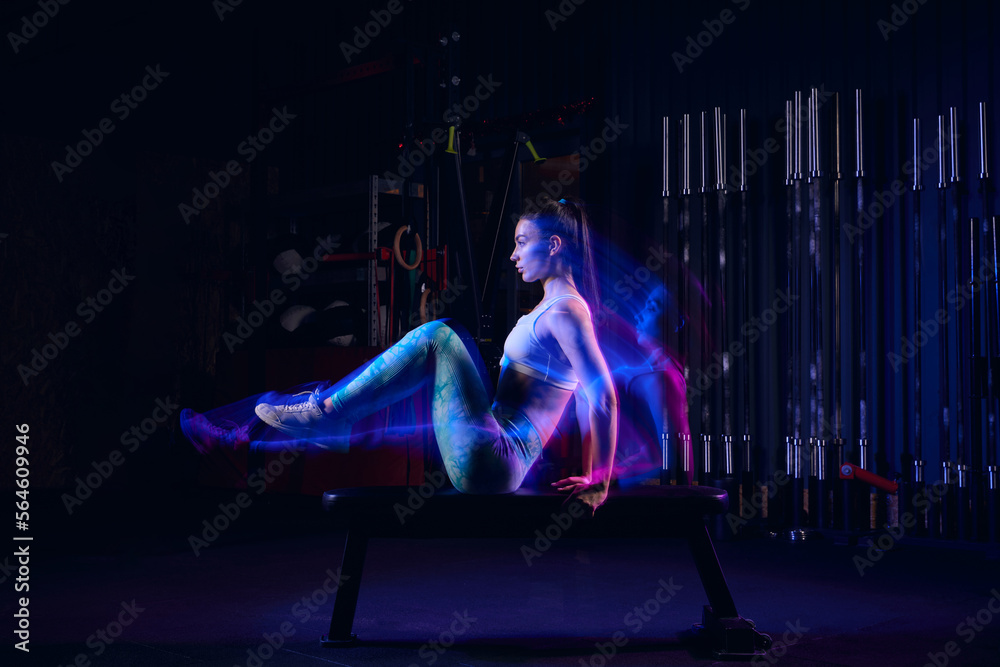 Young athletic woman wearing sports uniform and sneakers going in for sports, fitness training in modern gym. Dark background with mixed neon light