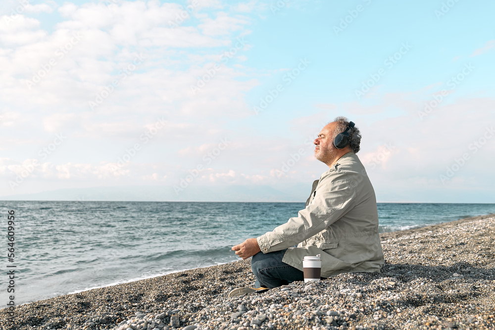 Happy middle-aged bearded man sitting in lotus pose on pebble winter beach, meditating and listening music or podcast from smartphone application in headphones. Relaxing outdoors. Happy middle-aged bearded man sitting in lotus pose on pebble winter beach, meditating and listening music or podcast from smartphone application in headphones. Relaxing outdoors.