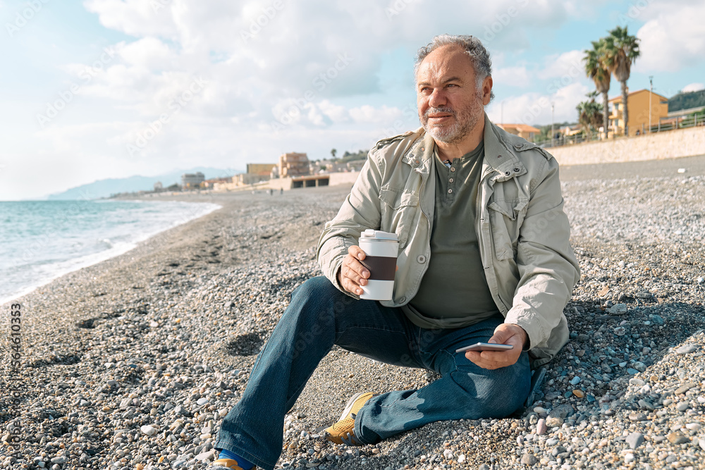 Happy middle-aged bearded man in casual clothes using smartphone and drinking coffee while while sitting on winter on spring beach. Relaxing outdoors.