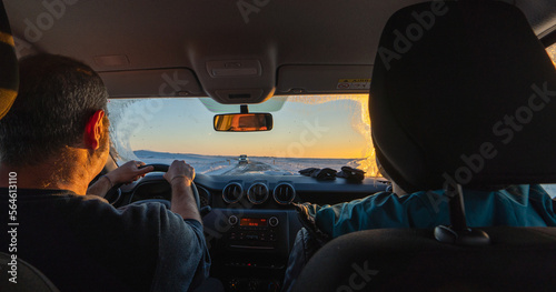 A middle-aged couple driving a 4x4 from inside the car at dawn on a totally snowy road with the sun rising on the horizon and the driver's gaze reflected in the rearview mirror