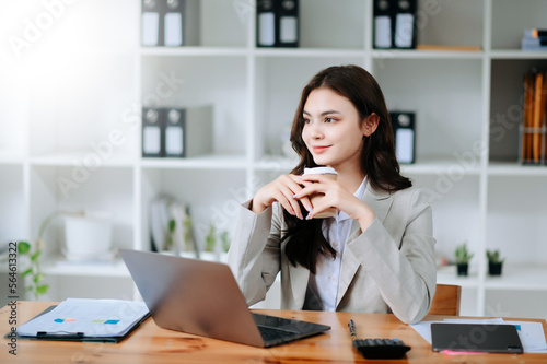 Confident beautiful Asian businesswoman typing laptop computer and digital tablet while holding coffee at office..