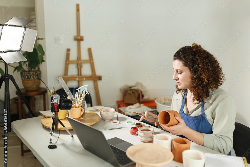 Females potter in blue apron making a vlog, recording online course, clay master class,lessons in her studion with earthenware.Using led lamp,studio equipment Females potter in blue apron making a vlog, recording online course, clay master class,lessons in her studion with earthenware.Using led lamp,studio equipment