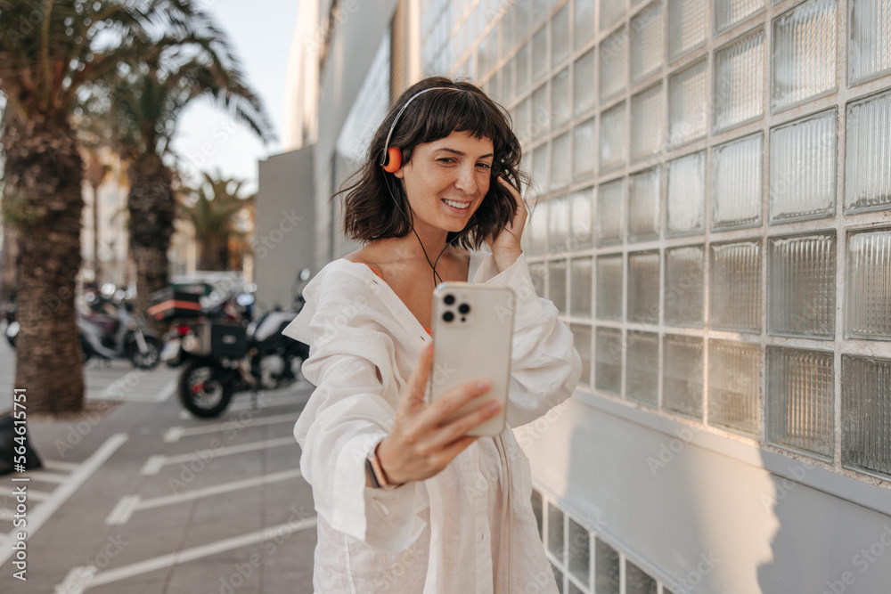 Charming fair-skinned young woman doing selfie in phone at the street. In headphones, look with smile on the street background. Positive emotion, technology concept 