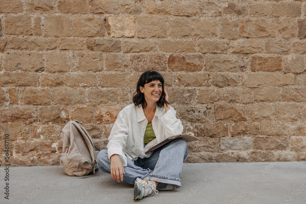 Caucasian smiling woman sitting on the floor with backpack and notebook. Wearing casual clothes, looking away, touching hair. Lifestyle concept Caucasian smiling woman sitting on the floor with backpack and notebook. Wearing casual clothes, looking away, touching hair. Lifestyle concept