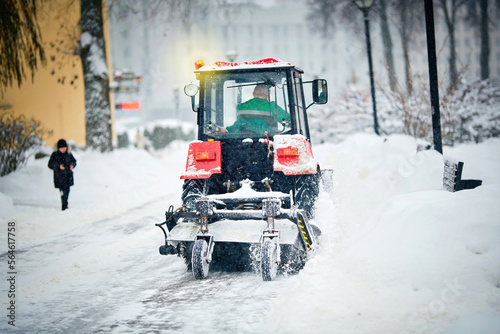 Tractor sweep, plow snow with rotating brush and snowplow from sidewalk. Snow plow vehicle remove snow and ice from road, winter road maintenance during blizzard. Tractor clear snowy street