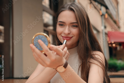Beautiful young woman with cosmetic pocket mirror applying lipstick outdoors