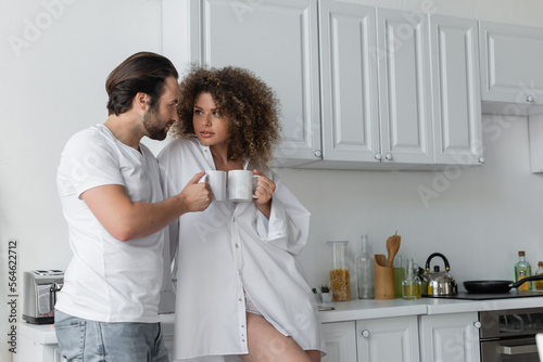 bearded man and curly woman holding cups of coffee in kitchen.