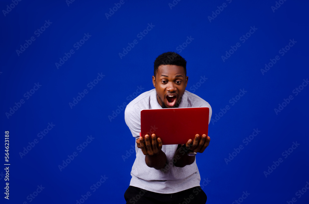 portrait of African man surprised with his mouth opened carrying a laptop isolated on a blue background portrait of African man surprised with his mouth opened carrying a laptop isolated on a blue background