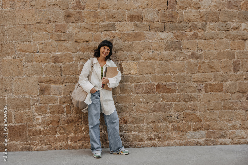 Full view of european beautiful woman holding notebooks with backpack. Smiling dark-haired student staying on the wall background. Lifestyle concept