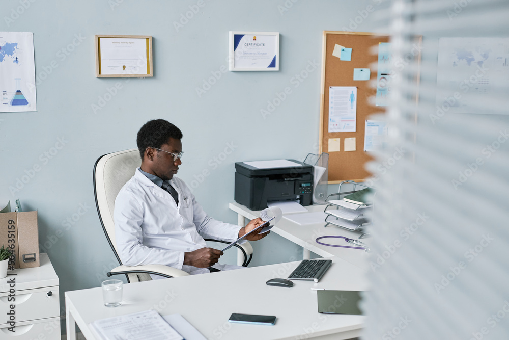 Wide angle young black doctor relaxing in chair at office reading documents
