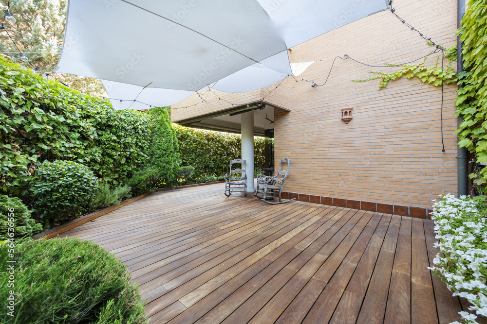 Terrace of a ground floor house with hedges and ornamental plants, slatted acacia hardwood floors twin wooden sunbeds and white awnings - obrazy, fototapety, plakaty 