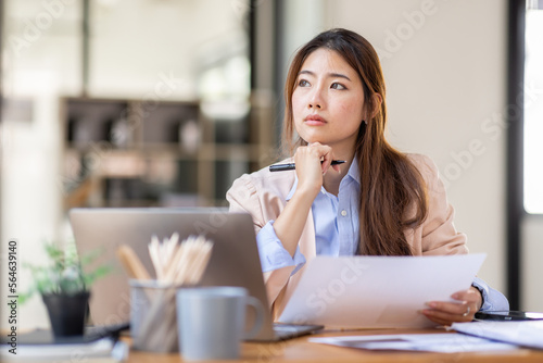 Image of young beautiful brooding Asian woman working with laptop while sitting at laptop in office, thinking of professional plan, project management, considering new business ideas.