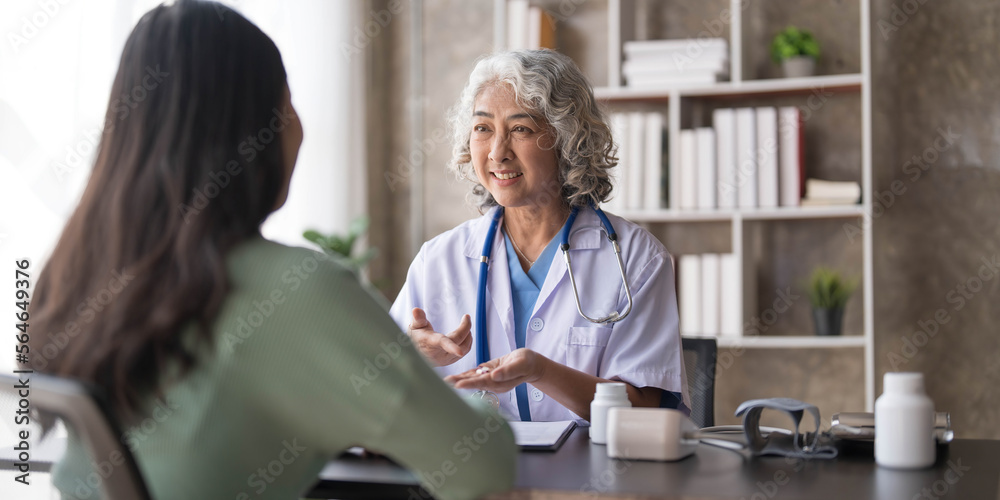 Fototapeta premium Woman senior doctor is Reading Medical History of Female Patient and Speaking with Her During Consultation in a Health Clinic. Physician in Lab Coat Sitting Behind a Laptop in Hospital Office.